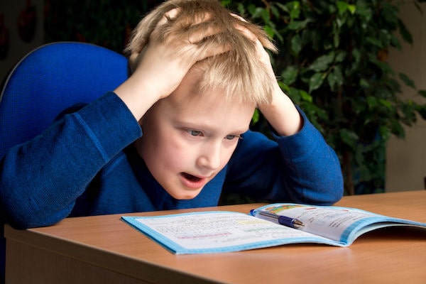 young boy looking at book on desk with hands on head looking frustrated