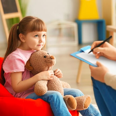 young child holding teddy bear sat opposite person holding clipboard